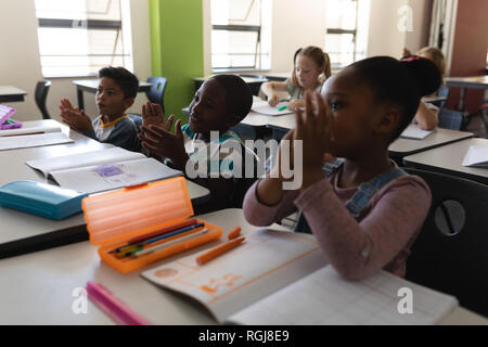 Vue latérale du schoolkids applaudir et sitting at desk in classroom Banque D'Images