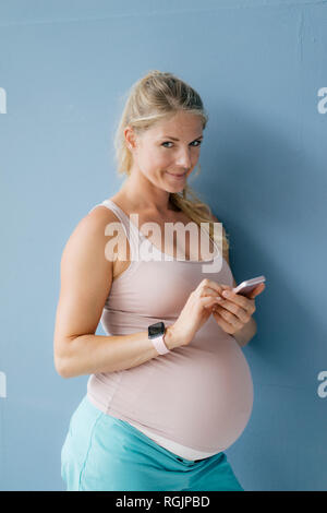 Portrait of smiling pregnant woman standing at blue wall using cell phone Banque D'Images