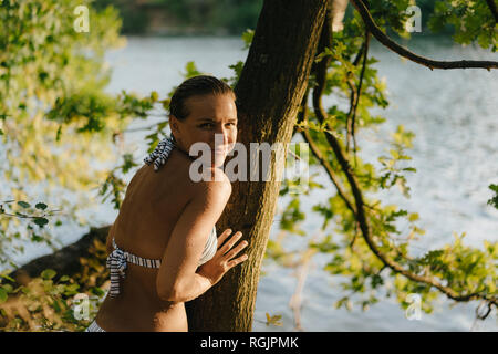 Portrait of smiling woman wearing a bikini au tronc de l'arbre au bord d'un lac Banque D'Images