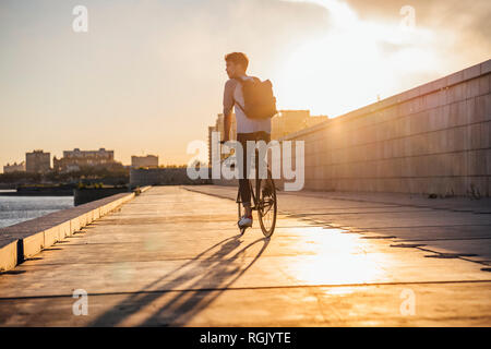 Jeune homme avec sac à dos équitation vélo sur la promenade du front de mer au bord de la rivière au coucher du soleil Banque D'Images
