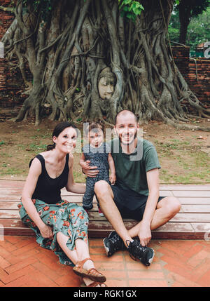 La Thaïlande, Ayutthaya, portrait of smiling family visiting le bouddha en tête entre les racines des arbres au Wat Mahathat Banque D'Images