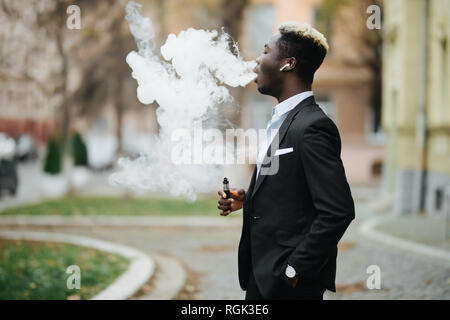 Homme africain dans un costume en vous promenant dans les rues de la ville avec la seine. Portrait d'un homme vaper avec nuage de fumée. Banque D'Images