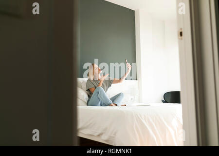 Jeune femme assise sur le lit en tenant un selfies Banque D'Images