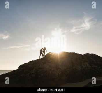 France, Bretagne, jeune couple d'escalade sur un rocher sur la plage au coucher du soleil Banque D'Images