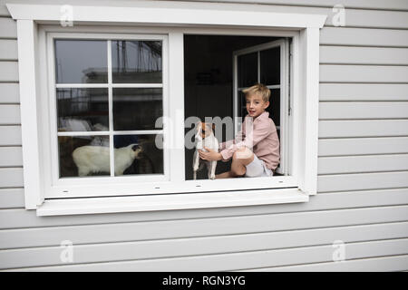 Portrait of boy sitting on window sill avec Jack Russel terrier et Siam chat hors de la fenêtre ouverte Banque D'Images