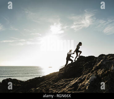 France, Bretagne, jeune couple d'escalade sur un rocher sur la plage au coucher du soleil Banque D'Images