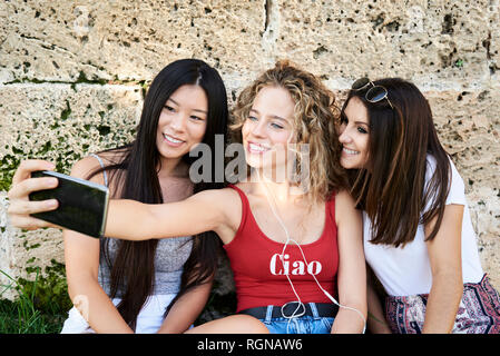 Trois jeunes femmes souriant assis à prendre un mur de pierre selfies Banque D'Images