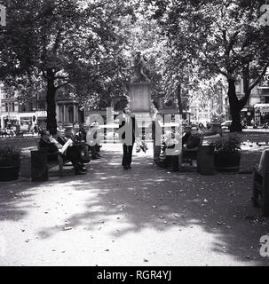 Années 1960, l'heure d'été et des gens assis sur des bancs dans le petit parc public ou d'un jardin à Leicester Square, dans le West End de Londres, Angleterre, Royaume-Uni. La statue de l'anglais paywright, William Shakespeare par Giovanni Fontana peut être vu dans la photo, une sculpture dans le centre de Leicester Square Gardens depuis 1874, un résultat d'améliorations apportées par Albert Grant qui a la place de cette même année. Banque D'Images