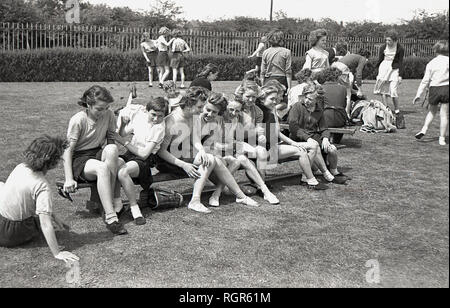 Années 1950, les filles s'amusant chatter avec les autres assis sur un banc à l'extérieur dans leur PE kit, prêt à participer à leur journée du sport scolaire, England, UK. Banque D'Images