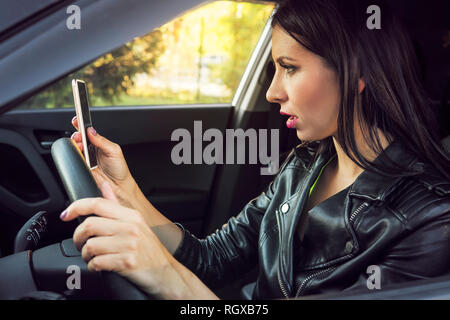 Brunette femme assise à l'intérieur de voiture avec un téléphone mobile dans la main texter au volant. Choqué girl contrôler son smartphone pas attention à road s Banque D'Images