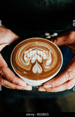 Une tasse de café avec un coeur de mousse latte art sur haut, être demande par mains masculines, couleurs marron chaleureux, tons de terre. Banque D'Images