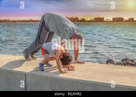 Le père et le jeune fils de démarrer le matin avec de l'exercice s'étend sur le bord de l'eau vire au mur comme le soleil se lève. Banque D'Images