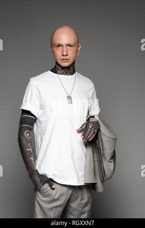 Bald grave tattooed man in white t-shirt tenue veste de costume et looking at camera isolé sur gray Banque D'Images