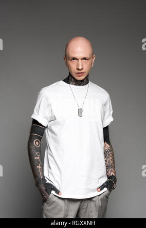 Handsome bald tattooed man in white t-shirt debout avec les mains dans les poches et looking at camera isolé sur gray Banque D'Images