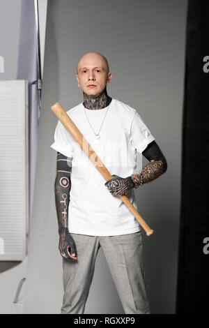 Tattooed man grave en blanc T-shirt holding baseball bat and looking at camera sur gris Banque D'Images