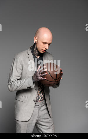 Bald grave tattooed man in suit holding basketball ball isolé sur gray Banque D'Images