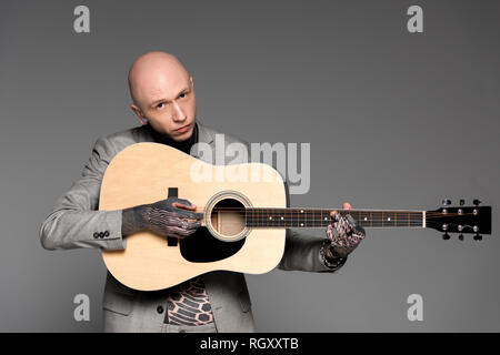 Handsome bald tattooed man in suit à jouer de la guitare et à la caméra à l'isolé sur gray Banque D'Images