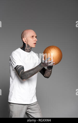 Bald tattooed man in white t-shirt jouer au basket-ball avec ballon d or isolé sur gray Banque D'Images