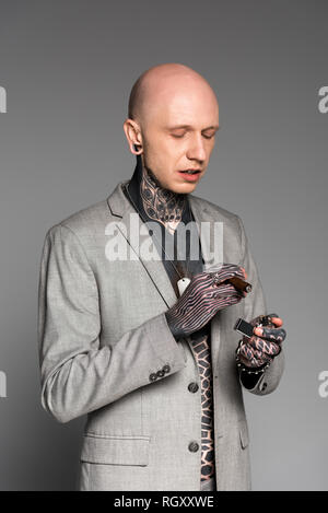 Bald tattooed man in suit jacket holding cigare et cigare isolé sur gray Banque D'Images