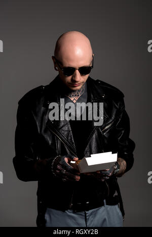 Tattooed man chauve en blouson de cuir et lunettes holding box avec de savoureux burger isolé sur gray Banque D'Images