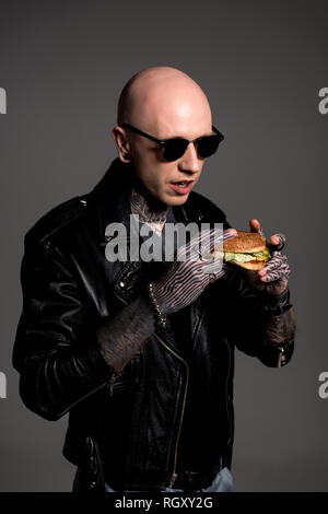 Tattooed man chauve en blouson de cuir et lunettes holding tasty burger isolé sur gray Banque D'Images