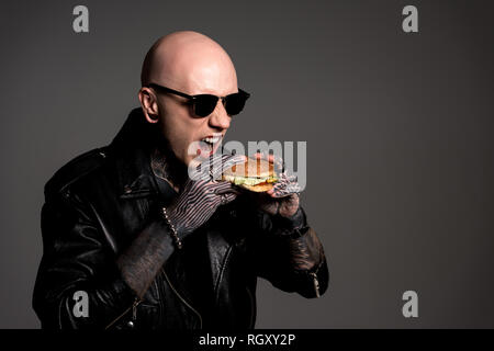 Tattooed man chauve en blouson de cuir et lunettes manger burger isolé sur gray Banque D'Images