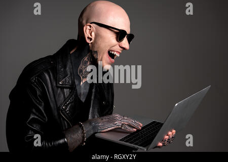 Tattooed man chauve en blouson de cuir et lunettes de soleil à l'aide d'ordinateur portable et rire isolé sur gray Banque D'Images