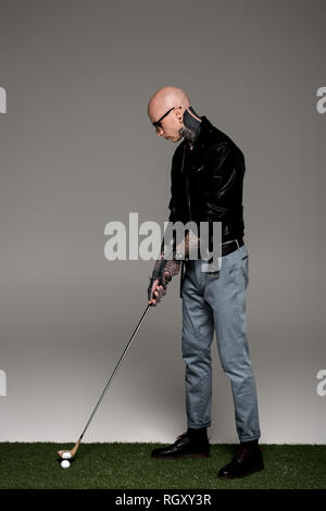 Tattooed man en jeans, blouson de cuir et lunettes de soleil en jouant au golf sur gris Banque D'Images