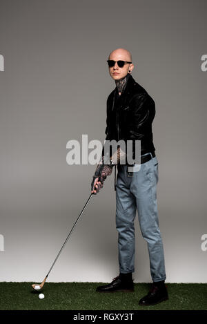Tattooed man élégant dans des lunettes de soleil et veste de cuir en jouant au golf sur gris Banque D'Images