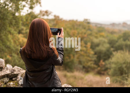 Une jeune femme aux cheveux rouge prend des photos avec son téléphone portable dans une forêt Banque D'Images