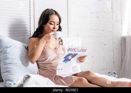 Young Asian woman boire du café et de la lecture de journaux d'entreprise, tout en restant au lit Banque D'Images