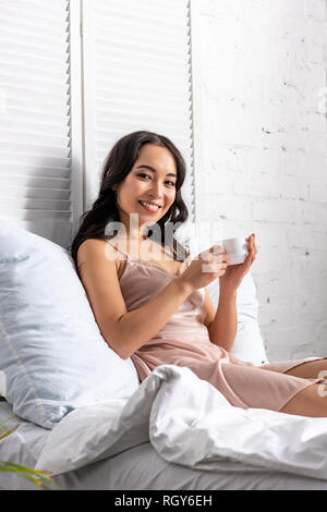 Jeune femme asiatique en nuisette élégante de boire du café tout en restant au lit et looking at camera Banque D'Images