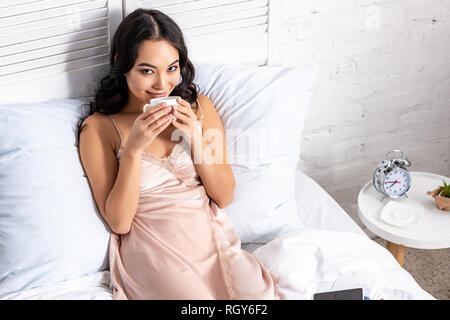 Beautifil asian woman in elegant nighty de boire du café tout en restant au lit et looking at camera Banque D'Images