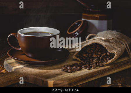 Tasse fumante de café avec café en grains déversés par un sac de toile sur une planche en bois dans un cadre rustique foncé Banque D'Images