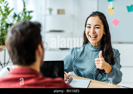 Smiling asian businesswoman sitting and showing thumb up sign pour in office Banque D'Images