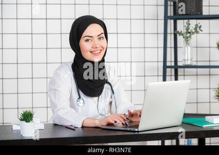 Femme scientifique voilée assise sur son bureau avec les doigts sur un clavier de son ordinateur portable, en souriant. Banque D'Images