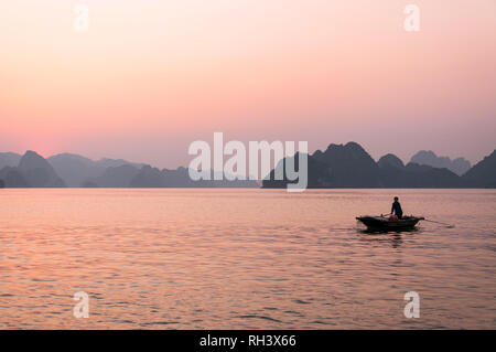 Une Vietnamienne aviron bateau en bois avec ses îles karstiques de calcaire en arrière-plan au coucher du soleil, la baie d'Halong, Vietnam Banque D'Images