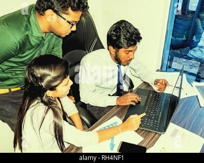 Équipe de deux jeunes hommes et une femme indienne dans l'office, au bureau avec ordinateur portable, des graphiques sur des feuilles c220300 smartphone. Banque D'Images