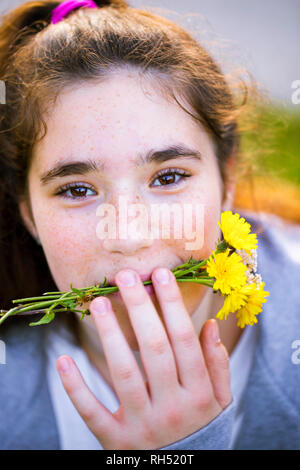 Close up portrait of a smiling teen fille avec de longs cheveux brun couchait et holding pissenlits jaunes dans sa bouche. Elle est mignonne et drôle. Banque D'Images