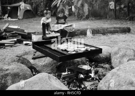 Un style australien bush barbecue avec cuisine des aliments sur un feu de camp avec deux enfants assis à l'arrière-plan, Finch Hatton, Queensland 4756, Australie Banque D'Images