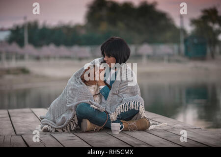 Portrait of beautiful young woman hugging her dog de la rivière. Femme heureuse avec son chien Basset Hound covered with blanket assis sur la jetée en bois Banque D'Images