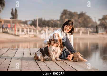 Portrait of beautiful young woman Playing with dog par la rivière. Happy woman sitting on the wooden pier avec son chien Basset Hound. Femme avec chiot. Banque D'Images