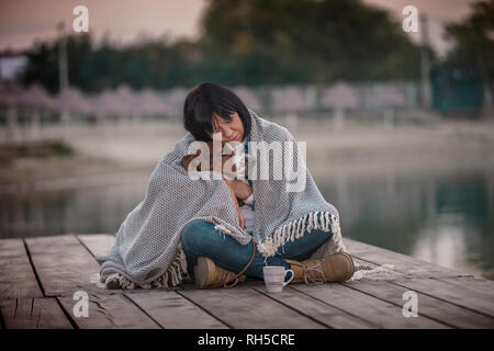 Portrait of beautiful young woman hugging her dog de la rivière. Femme heureuse avec son chien Basset Hound covered with blanket assis sur la jetée en bois Banque D'Images