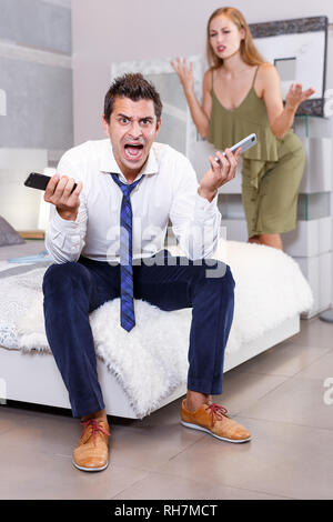 Portrait de crier en colère homme assis avec deux téléphones portables sur lit dans chambre famille Banque D'Images