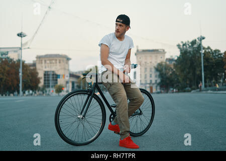 Portrait de plein air d'un jeune homme moderne dans la rue, assis sur le vélo. Un jeune homme portant un pantalon athlétique kaki, blanc T-shirt, sac à dos à roulettes rouge sneakers et Banque D'Images