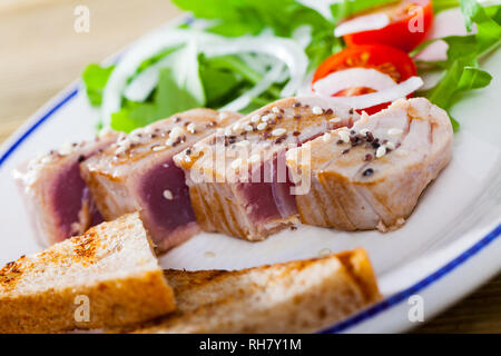 Délicieux thon frit légèrement saupoudré de sésame servi sur une assiette avec du pain grillé, tomates fraîches et vertes. Banque D'Images