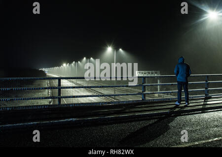 Le dirigeant d'une silhouette encapuchonnée debout sur le pont à la recherche vers le bas sur une autoroute vide la nuit, pendant une tempête de neige. Banque D'Images