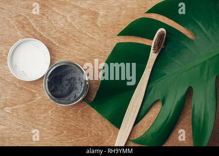 Concept zéro déchet. Dentifrice naturel du charbon activé en bocal de verre et brosse à dents en bambou sur fond de bois avec des feuilles vert monstera. Fr en plastique Banque D'Images