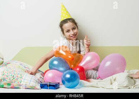 L'anniversaire d'une adolescente est de 10-11 ans. Une fille dans un chapeau de fête avec un cadeau se trouve sur le lit dans une chambre d'enfants, des ballons d'arrière-plan Banque D'Images