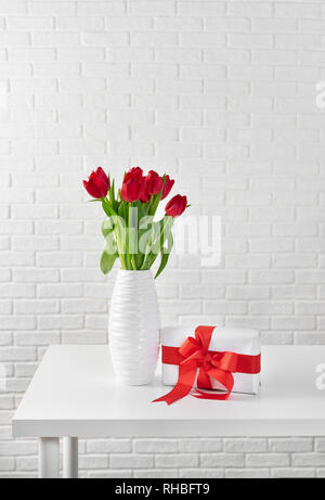 Tulipes rouges dans vase blanc à côté d'une boîte cadeau avec ruban rouge contre le mur brocks blanc Banque D'Images